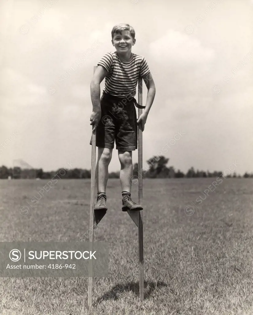1930S 1940S Smiling Happy Boy Wearing Striped Shirt & Short Pants Walking On Pair Of Stilts Looking At Camera