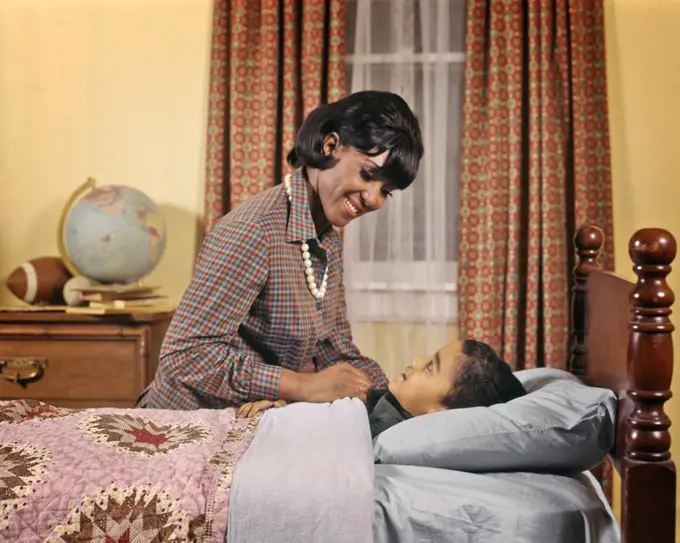 1970s SMILING AFRICAN AMERICAN WOMAN MOTHER TUCKING GIRL DAUGHTER INTO BED AT NIGHT