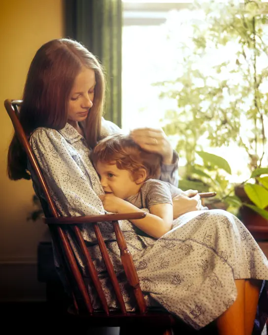 1970s WOMAN SITTING IN ROCKING CHAIR COMFORTING BOY TODDLER STANDING NEXT TO HER BY HOUSEPLANT FILLED WINDOW