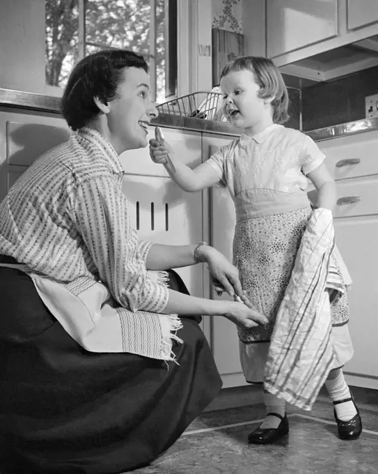 1950s LITTLE GIRL AND MOTHER IN KITCHEN BOTH WEARING APRONS GIRL HOLDING DISH TOWEL GESTURING TO MOTHER