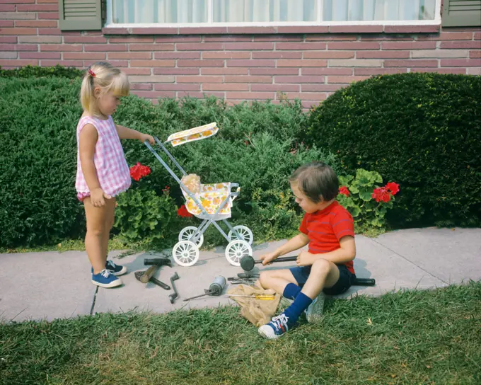 1970s LITTLE GIRL PUSHING TOY BABY CARRIAGE AND BOY PLAYING WITH TOOLS ON SIDEWALK GENDER SPECIFIC ROLE PLAY