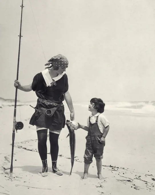 1920s WOMAN MOTHER HOLDING SURF CASTING FISHING POLE CARRYING FISH CATCH WITH BOY SON WALKING ON  OCEAN SEASHORE BEACH SAND