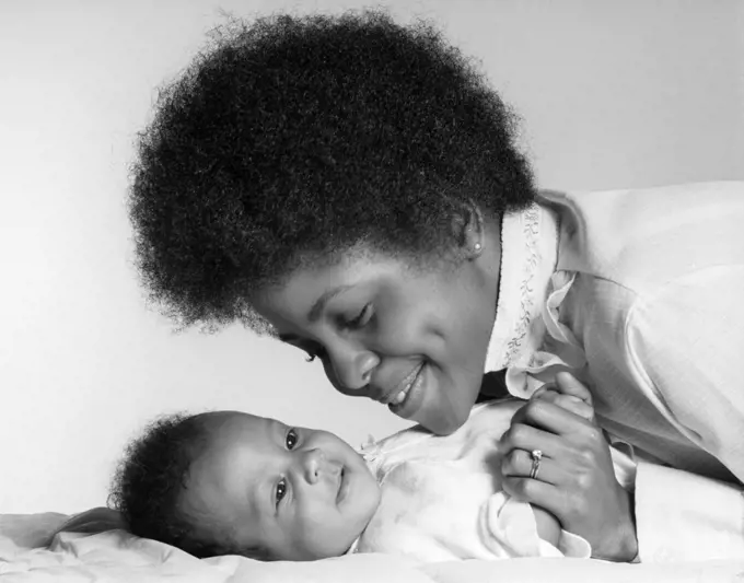 1970s SMILING AFRICAN AMERICAN MOTHER HOLDING HAND AND LOOKING AT HAPPY BABY DAUGHTER LYING ON HER BACK