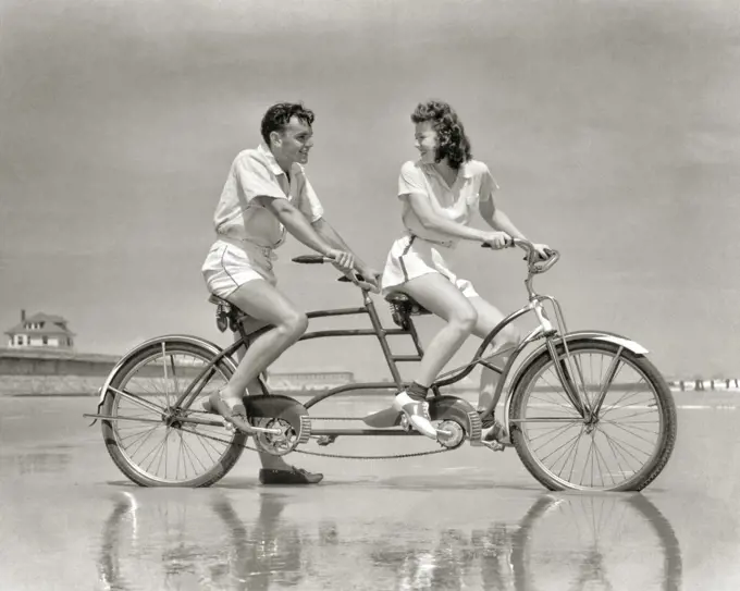 1940s YOUNG COUPLE IN WHITE SPORTS CLOTHES RIDING TANDEM BICYCLE AT SEASHORE ON WET BEACH WOMAN SMILING LOOKING BACK AT MAN