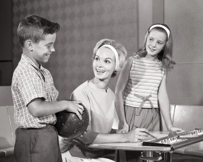 1960s Smiling mom keeping score at bowling alley with daughter and son who is holding a bowling ball
