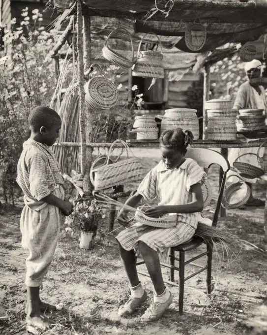 1940s AFRICAN AMERICAN BOY STANDING WATCHING GIRL WEAVING BASKET AT STAND WITH SEVERAL BASKETS FOR SALE SOUTH CAROLINA USA