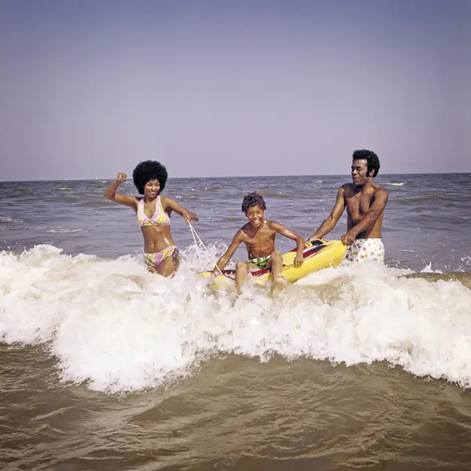 1970s AFRICAN-AMERICAN FAMILY JUMPING IN OCEAN SEASHORE SURF MOTHER AND FATHER WITH SON ON INFLATABLE RAFT