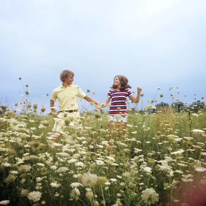 1970s TEENAGE COUPLE HOLDING HANDS RUNNING THROUGH A FIELD OF QUEEN ANNES LACE SMILING WEARING SUMMER CLOTHES