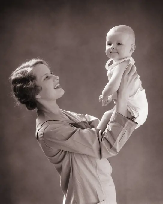 1930s SMILING MOTHER LIFTING UP BALD BABY BOY WITH FUNNY FACIAL EXPRESSION WEARING CLOTH DIAPER