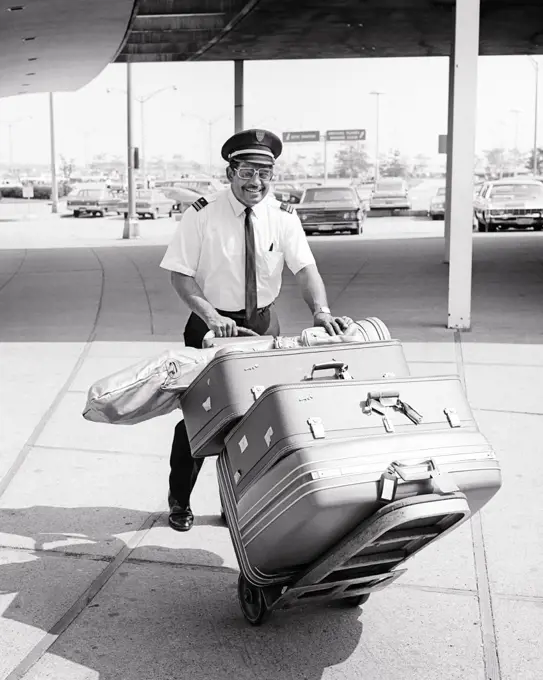 1960s SMILING AFRICAN-AMERICAN MAN AIRPORT PORTER PUSHING CART FULL OF LUGGAGE AT CURBSIDE