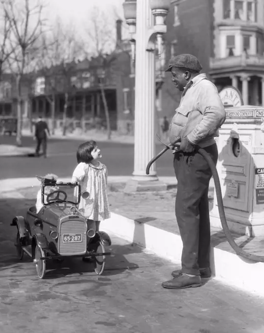 1920S Smiling Little Girl Standing Beside Her Pedal Car Asking For Gasoline From African American Gas Pump Attendant