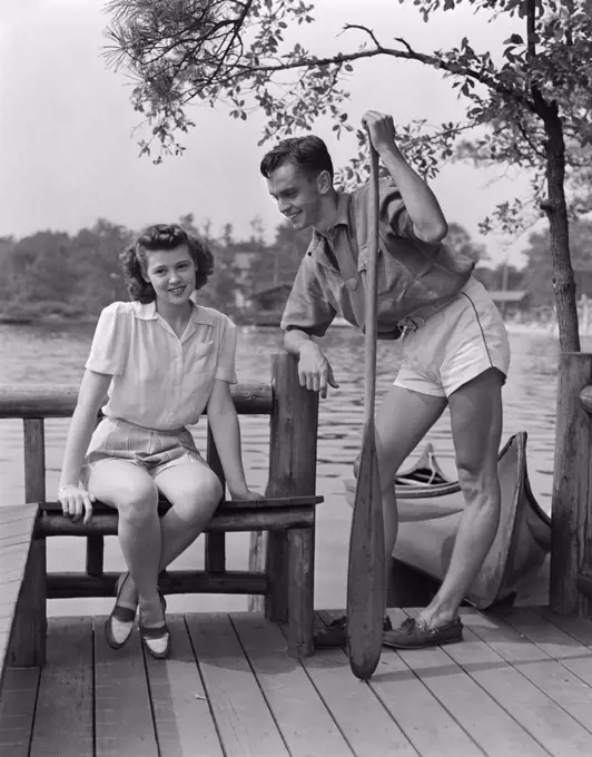 1940S Man And Woman On Dock By Lake Woman Sitting Man Standing Holding Oar Canoe In Background Summer Flirt