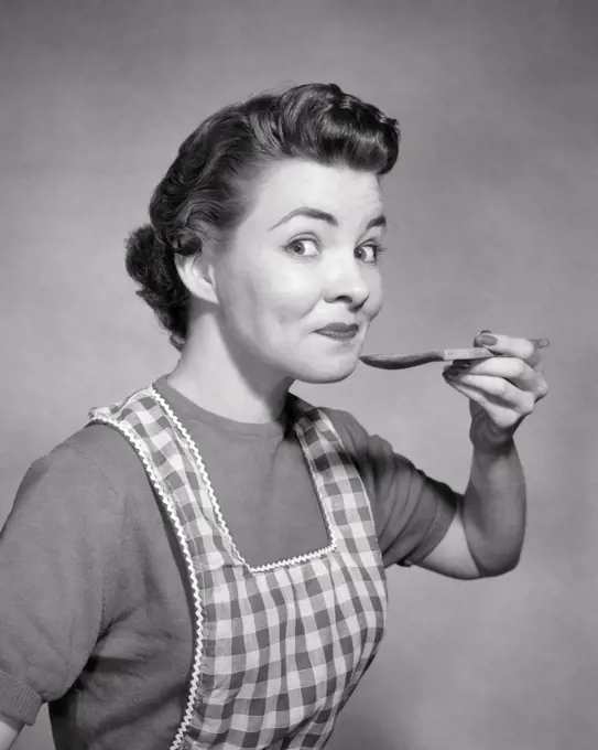 1950s SMILING WOMAN HOUSEWIFE WEARING CHECKERED APRON LOOKING AT CAMERA WITH RAISED EYEBROWS TASTING FROM WOODEN KITCHEN SPOON