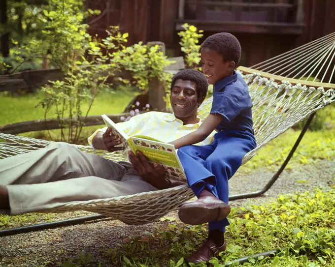 1970s SMILING AFRICAN-AMERICAN MAN IN A BACKYARD HAMMOCK READING A BOOK WITH HIS SON