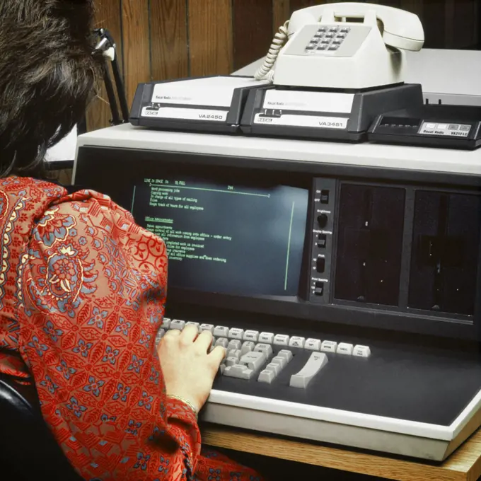 1980s BACK OF ANONYMOUS WOMAN WEARING RED PAISLEY DRESS WORKING ON EARLY COMPUTER TERMINAL KEYBOARD WITH DIAL-UP TELEPHONE MODEM