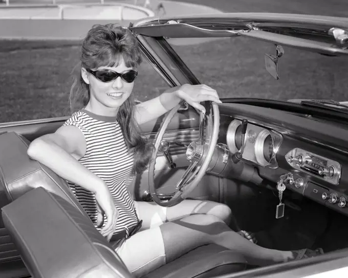 1960s SMILING  PRETTY TEEN GIRL WEARING SUNGLASSES LOOKING AT CAMERA SITTING AT STEERING WHEEL IN CONVERTIBLE CAR DRIVERS SEAT