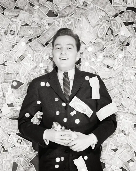 1950s SMILING  MAN LYING ON BACK ON A PILE OF MONEY COINS AND BILLS LOOKING AT CAMERA WINNER LUCKY LOTTERY PRIZE MONEY