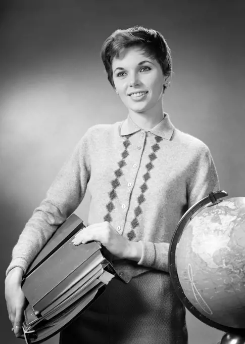 1950s 1960s  BRUNETTE WOMAN TEACHER CARRYING CLASS MATERIALS  BOOKS NOTEBOOKS SMILING STANDING BY GLOBE WEARING SWEATER