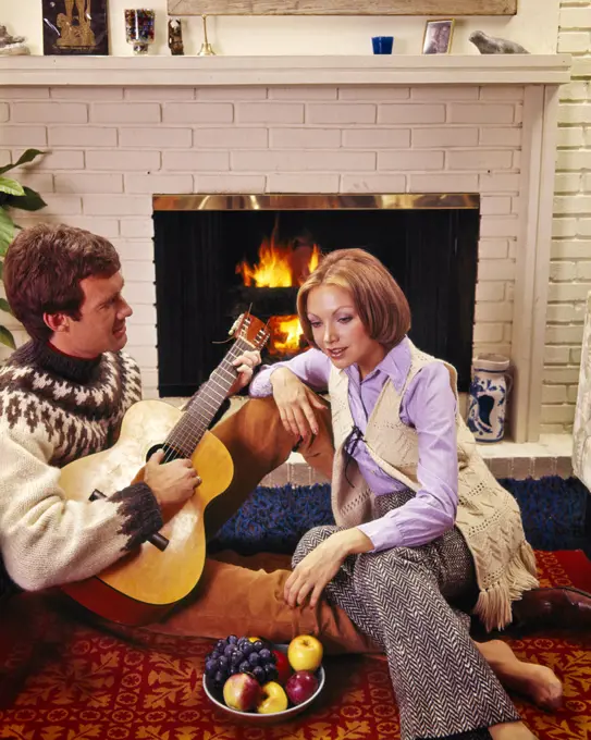 1960s 1970s COUPLE SITTING TOGETHER BY BRICK FIREPLACE MAN PLAYING GUITAR BAREFOOT WOMAN WEARING BELLBOTTOMS LISTENING ENTRANCED