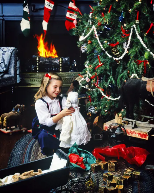 1970s 1980s SMILING BLOND GIRL HOLDING ANTIQUE DOLL SITTING BY TRADITIONAL CHRISTMAS TREE AND FIREPLACE ANTIQUE TOYS ALL AROUND