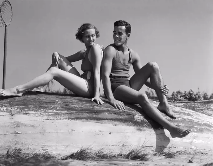 1930S Couple Man Woman Smiling Wearing Bathing Suits Sitting Back To Back On Overturned Boat  