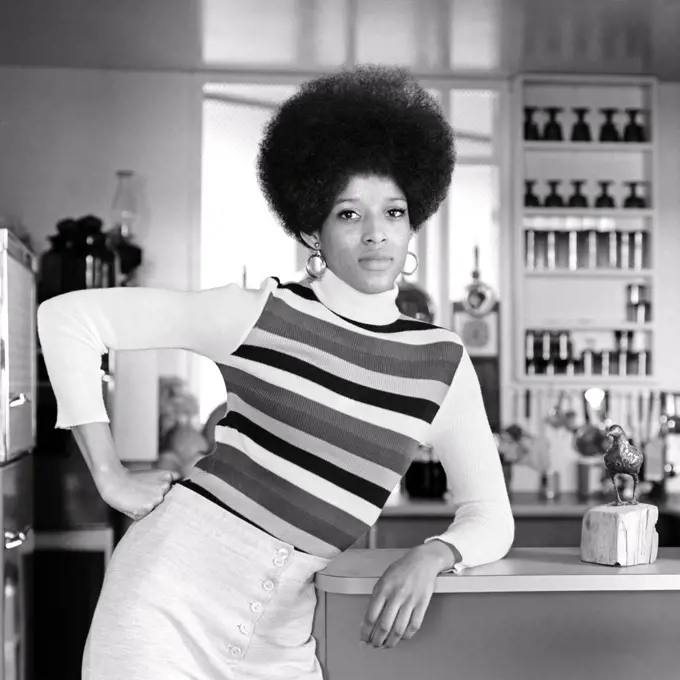 1960s 1970s AFRICAN-AMERICAN WOMAN TALL AFRO HAIRSTYLE STRIPED SHIRT LEANING ON RESTAURANT COUNTER HAND ON HIP LOOKING AT CAMERA