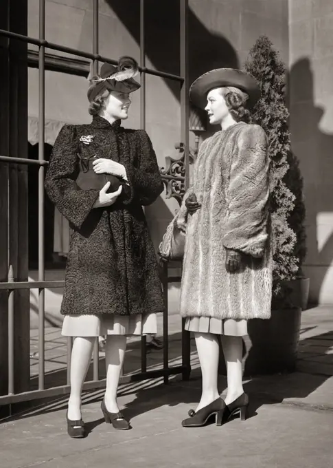 1930s 1940s TWO FASHIONABLE WOMEN WEARING HATS FUR COATS GLOVES STANDING MEETING TALKING TOGETHER SHOPPING OUTSIDE UPSCALE STORE