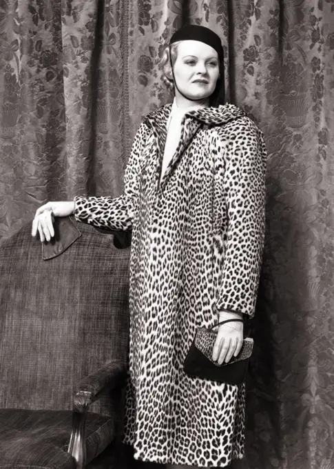 1930s 1938 FASHIONABLE WOMAN WEARING LEOPARD SKIN COAT STANDING BY CHAIR HOLDING ALLIGATOR HAND BAG WEARING LEATHER GLOVES