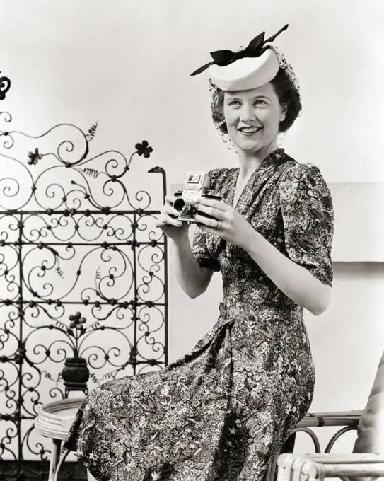 1940s SMILING WOMAN PHOTOGRAPHER LOOKING AT CAMERA HOLDING REFLEX CAMERA WEARING PRINT DRESS SILLY HAT SEATED ON OUTDOOR PATIO