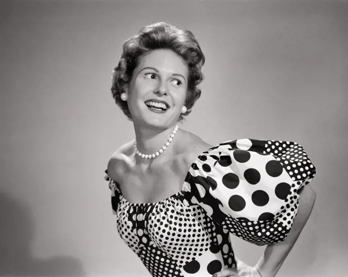 1950s SMILING WOMAN LOOKING OVER HER SHOULDER POSING WEARING LOW CUT SCOOP NECKLINE DRESS WITH POLKA DOTS AND PUFFY SLEEVES