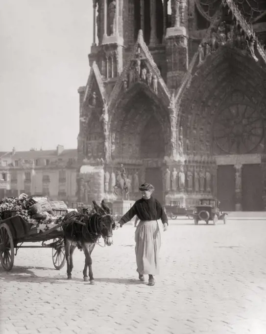 1920s ELDERLY WOMAN LEADING DONKEY CART LADEN WITH FOOD FRUITS AND VEGETABLES PAST THE FRONT OF THE CATHEDRAL RHEIMS FRANCE