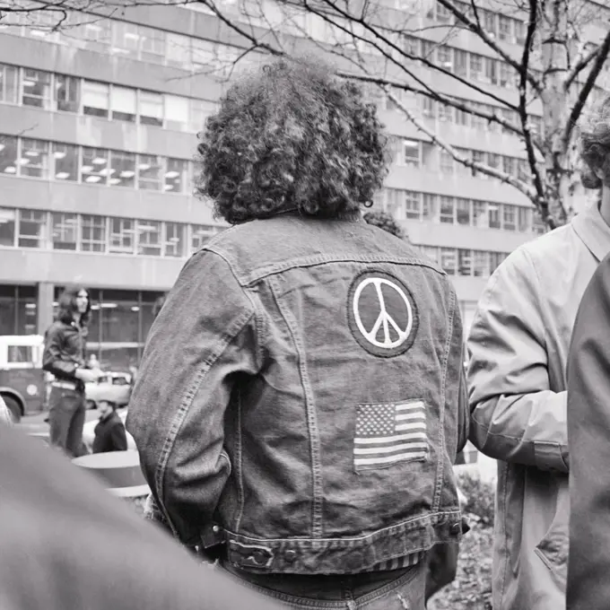 1960s 1970s ANONYMOUS MALE HIPPIE BACK VIEW LONG CURLY HAIR BLUE JEAN DENIM JACKET AMERICAN FLAG AND PEACE SYMBOL PATCHES