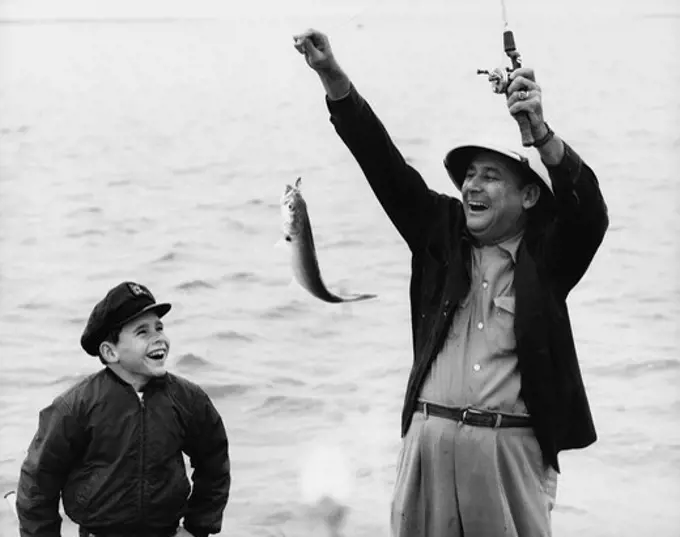 1950s 1960s BOY SON FISHING WITH MAN FATHER OR GRANDFATHER HOLDING UP CAUGHT FISH ON LINE LAUGHING HAVING FUN