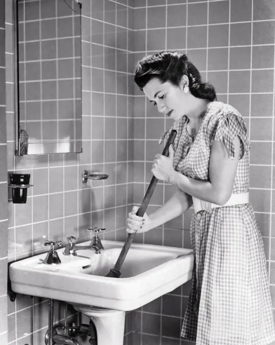 1940s ANNOYED EXASPERATED BRUNETTE WOMAN HOUSEWIFE IN THE BATHROOM USING A HAND PLUNGER TO CLEAR A CLOGGED SINK DRAIN