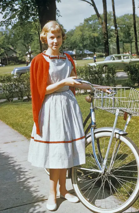 1950s BLOND TEENAGE GIRL WEARING DRESS AND RED CARDIGAN SWEATER STANDING WITH BICYCLE LOOKING AT CAMERA