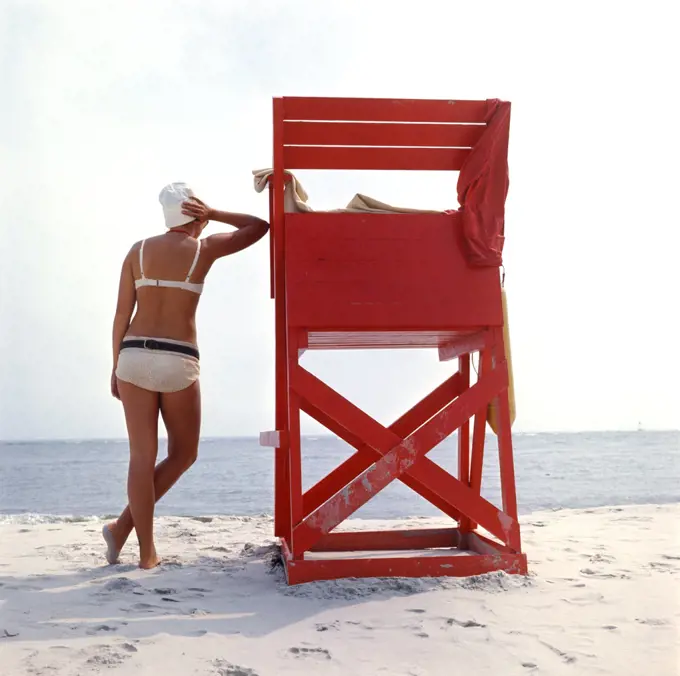 1980s REAR VIEW ANONYMOUS WOMAN IN WHITE BIKINI LEANING AGAINST LIFEGUARD STATION BEACH CHAIR BARNEGAT NEW JERSEY USA
