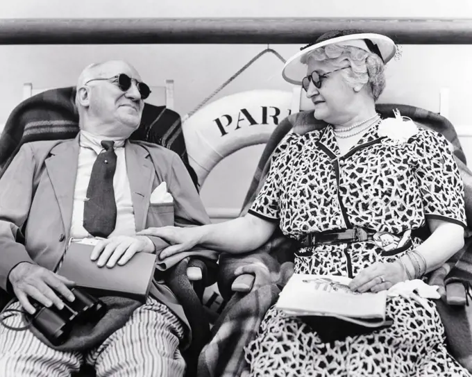 1930s SENIOR RETIRED COUPLE FASHIONABLY DRESSED SITTING IN DECK CHAIRS SIDE BY SIDE ON A CRUISE SHIP BOTH WEARING SUNGLASSES