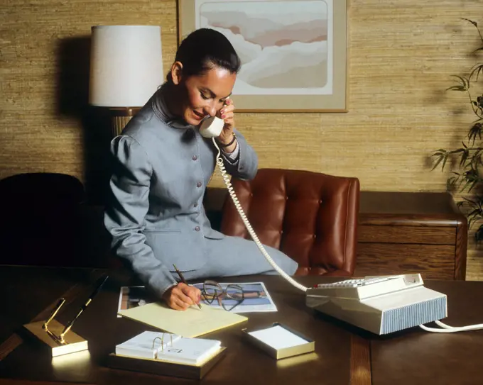 1980s SMILING BUSINESS WOMAN IN OFFICE SITTING ON HER DESK TALKING ON TELEPHONE WRITING NOTES