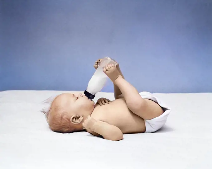 1940s BABY LYING ON BACK DRINKING FROM MILK BOTTLE HELD WITH FEET