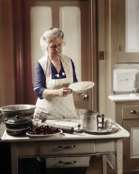 1920s 1930s SENIOR WOMAN GRANDMOTHER WEARING APRON CRIMPING CRUST MAKING A CHERRY PIE IN KITCHEN
