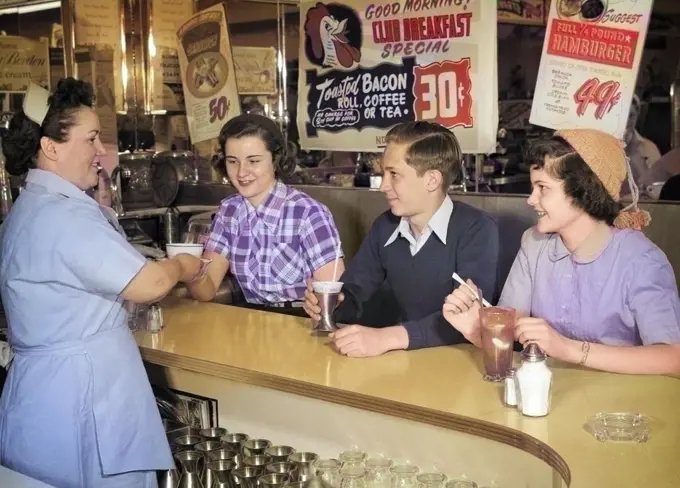1950s ANONYMOUS SMILING WOMAN WAITRESS SERVING TWO TEENAGE GIRLS AND ONE BOY AT DINER LUNCH SODA FOUNTAIN COUNTER
