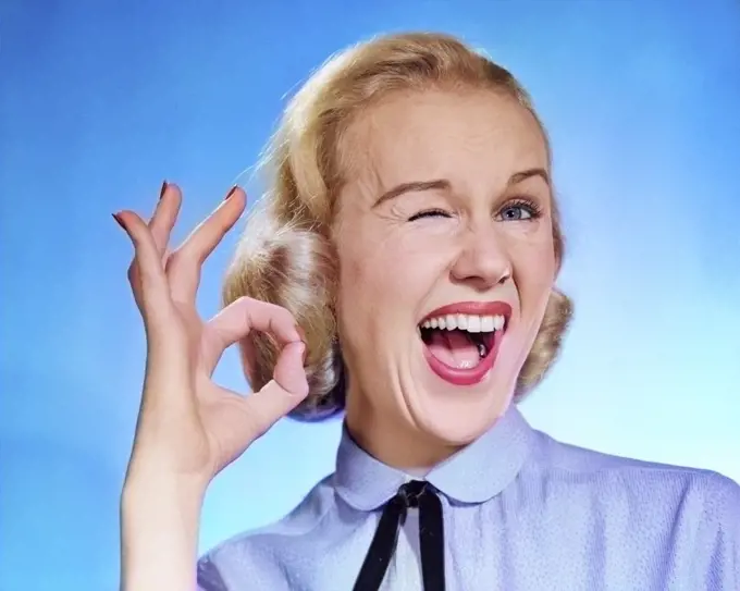 1950s ENTHUSIASTIC BLOND WOMAN MOUTH OPEN WINKING EYE LOOKING AT CAMERA AND MAKING OK SIGN WITH THUMB AND FINGER