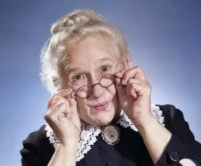 1950s PORTRAIT OF SMILING PRISSY OLD GRAY HAIR LADY HOLDING ADJUSTING HER ANTIQUE WIRE FRAME GLASSES LOOKING AT CAMERA POINTEDLY