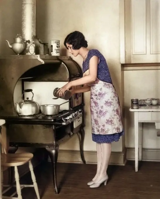 1920s HOUSEWIFE AT GAS STOVE AND OVEN COOKING IN KITCHEN