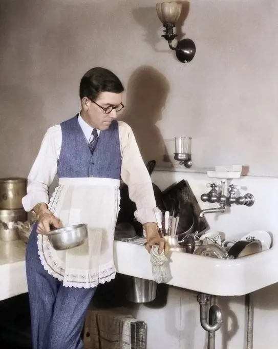 1920s 1930s PERPLEXED MAN IN APRON LEANING ON SINK FULL OF DIRTY DISHES RAG IN ONE HAND POT IN OTHER