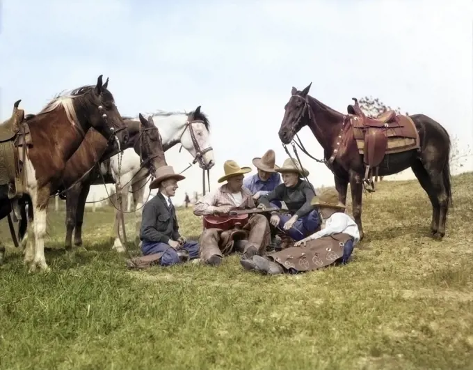 1920s 1930s GROUP OF FIVE SINGING COWBOYS SITTING ON GROUND SURROUNDED BY THEIR HORSES WHILE ONE PLAYS A GUITAR