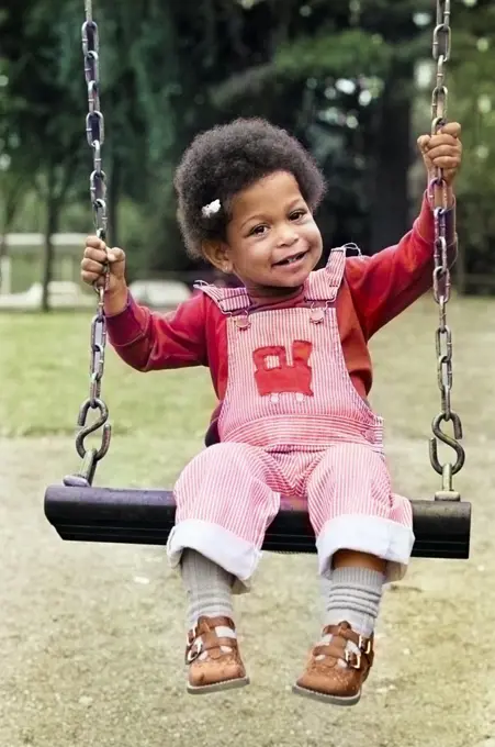 1970s 1980s CUTE CHARMING AFRICAN AMERICAN GIRL IN OVERALLS SITTING ON PLAYGROUND SWING LOOKING AT CAMERA