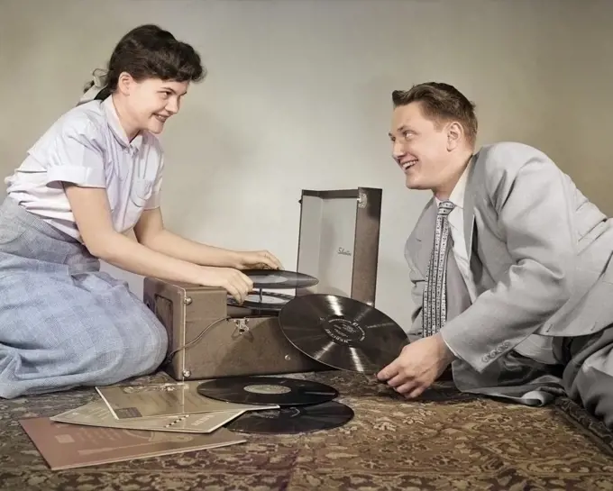1950s TEENAGE SMILING COUPLE BOY AND GIRL SITTING ON FLOOR PLAYING VINYL MUSIC RECORDS ON PORTABLE PHONOGRAPH