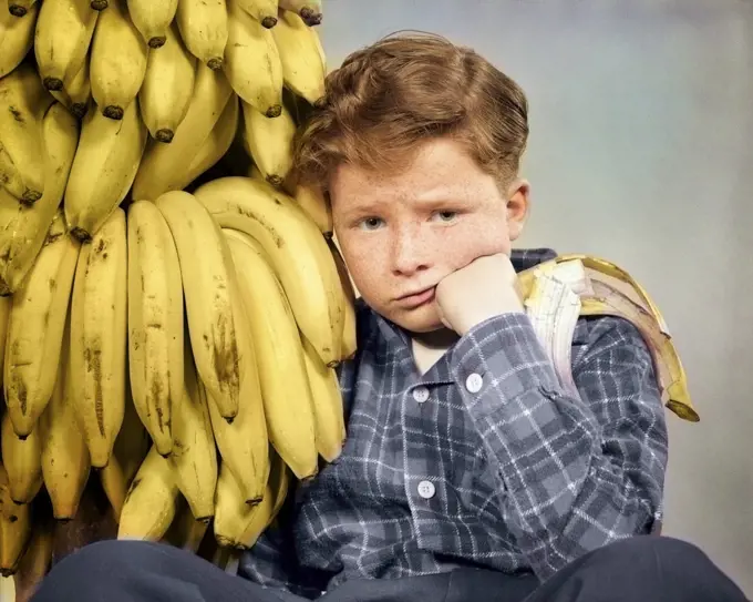 1930s 1940s DISTRESSED FRECKLE FACED BOY WITH STOMACH ACHE LOOKING AT CAMERA SITTING BY BUNCH OF BANANAS ONE HALF EATEN IN HAND