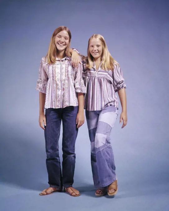 1970s TWO TEENAGE GIRLS STANDING LEANING ON EACH OTHER SMILING WEARING BELL-BOTTOM DENIM AND STRIPED SHIRTS LOOKING AT CAMERA